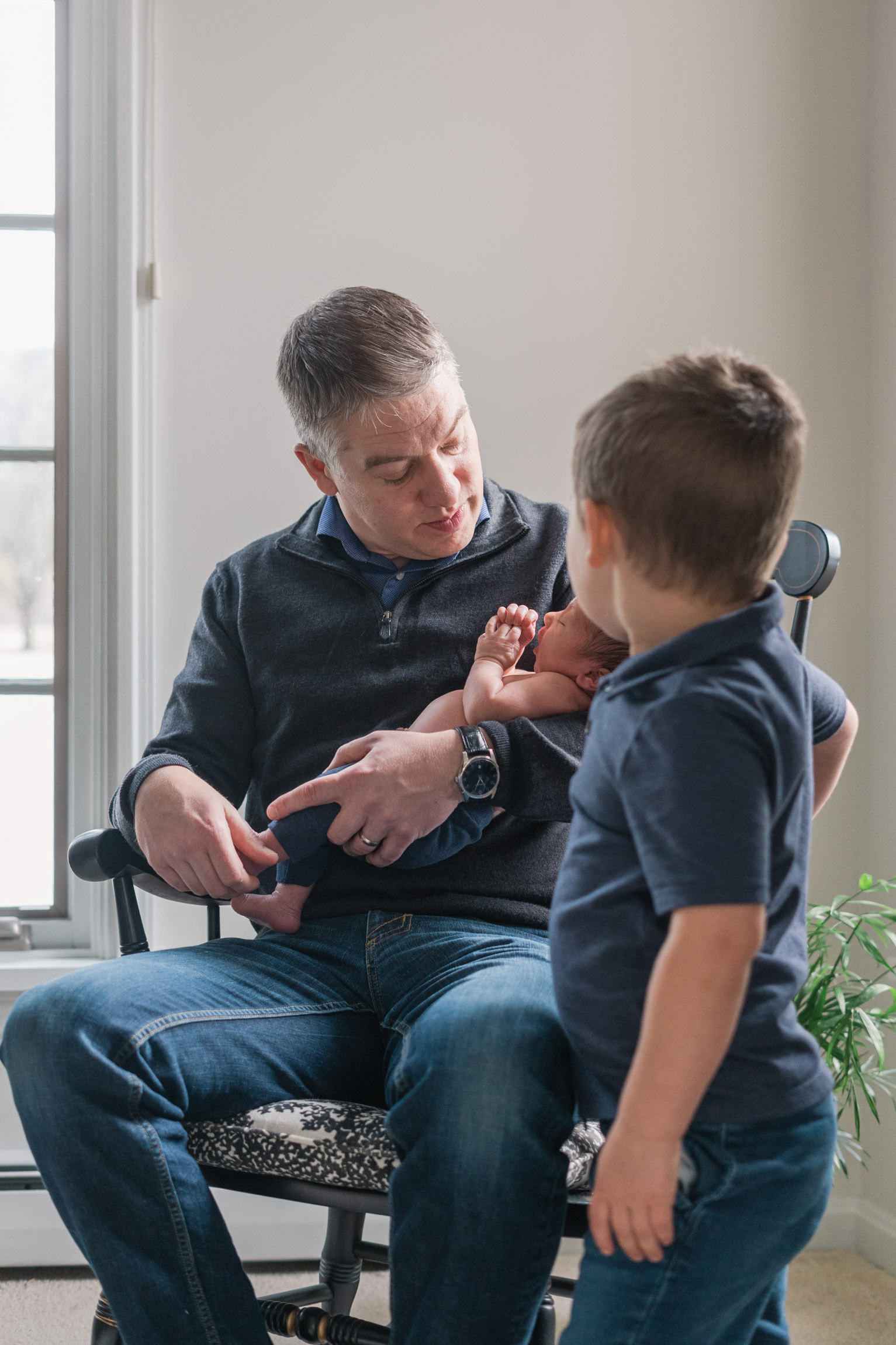 father holding newborn in rocking chair, other son looking on