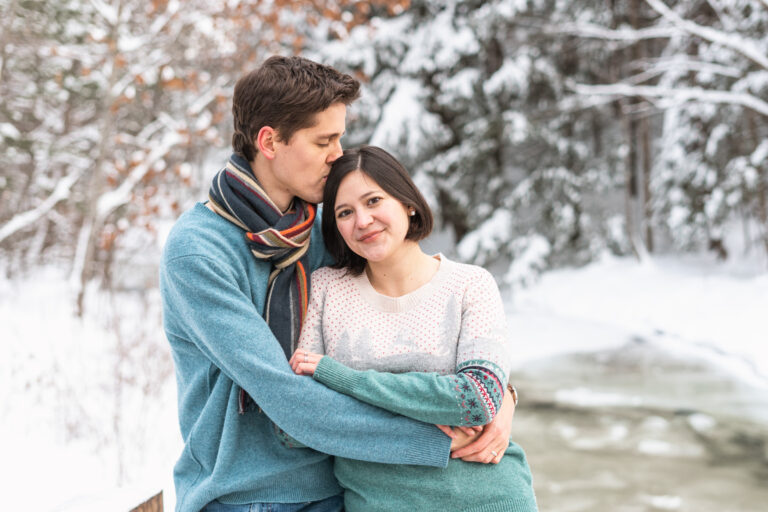 husband kissing top of wife's head with snowy river in background