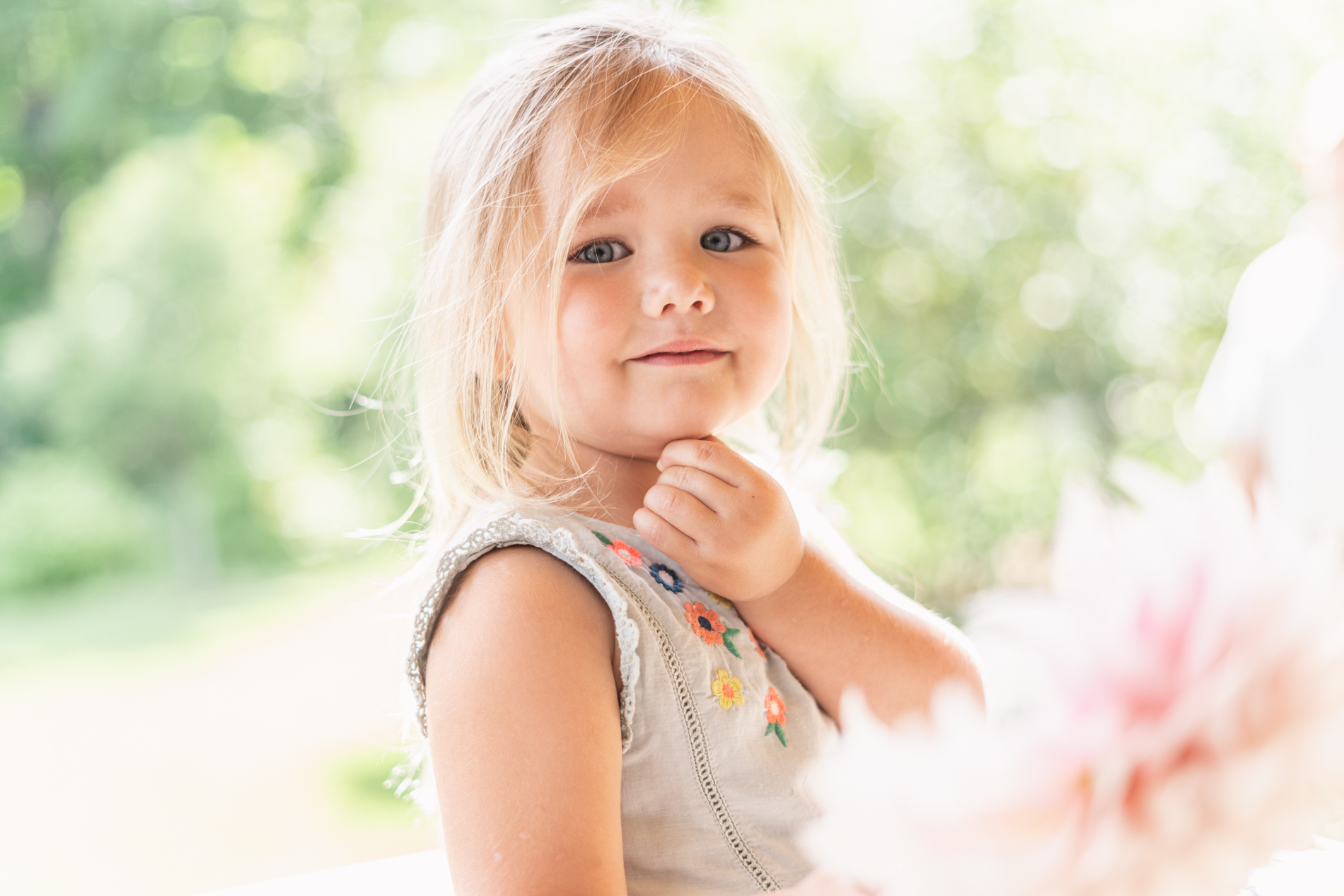 yung girl looking at camera with pink dahlia in foreground and green foliage in background