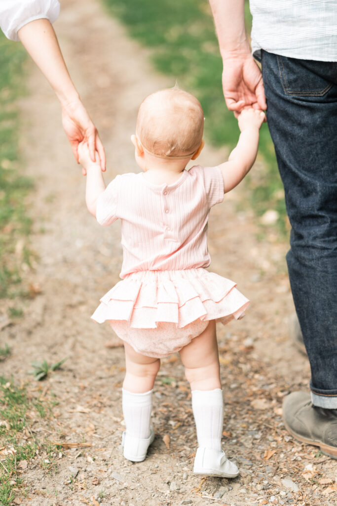 baby girl in pink ruffled outfit, walking away holding parents hands