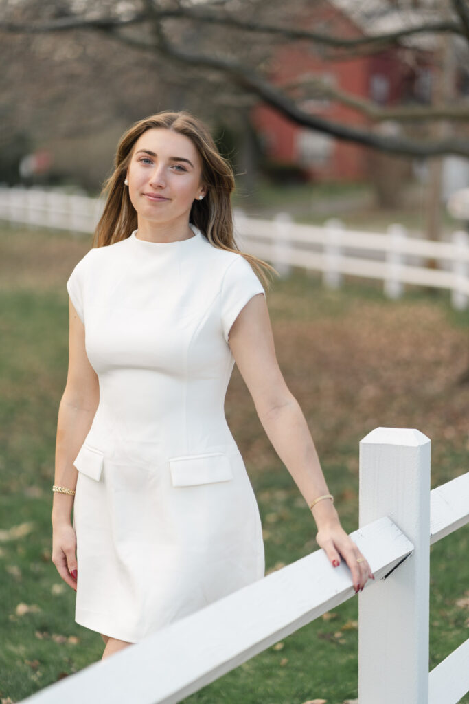 mid length portrait of female grad in white dress walking along fence near norwich university