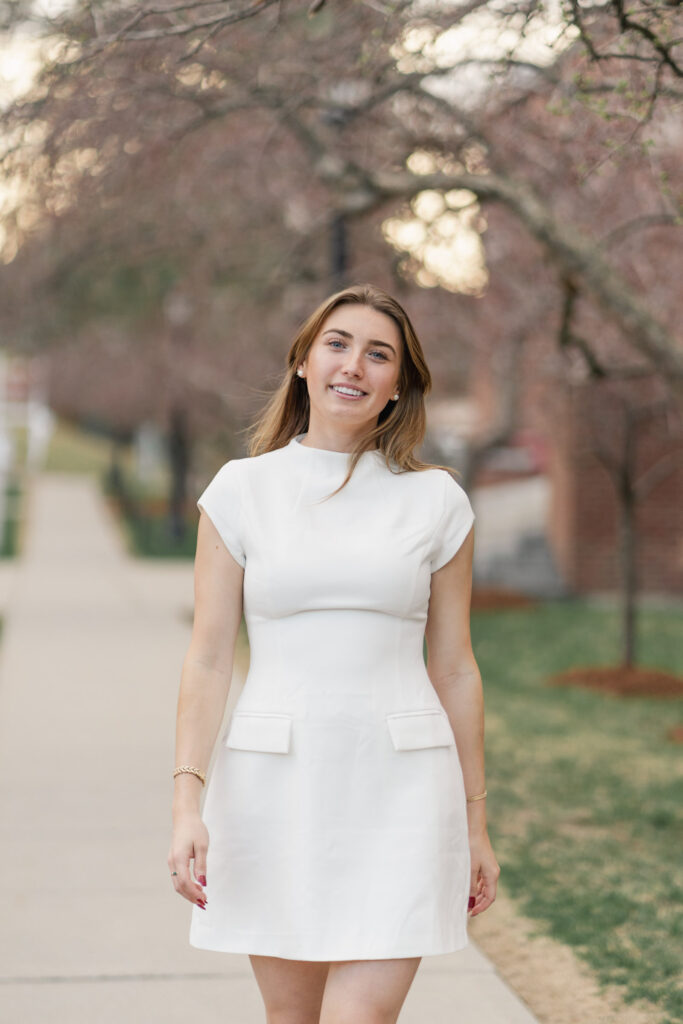 mid length portrait of female grad in white dress walking down backlit sidewalk near norwich university