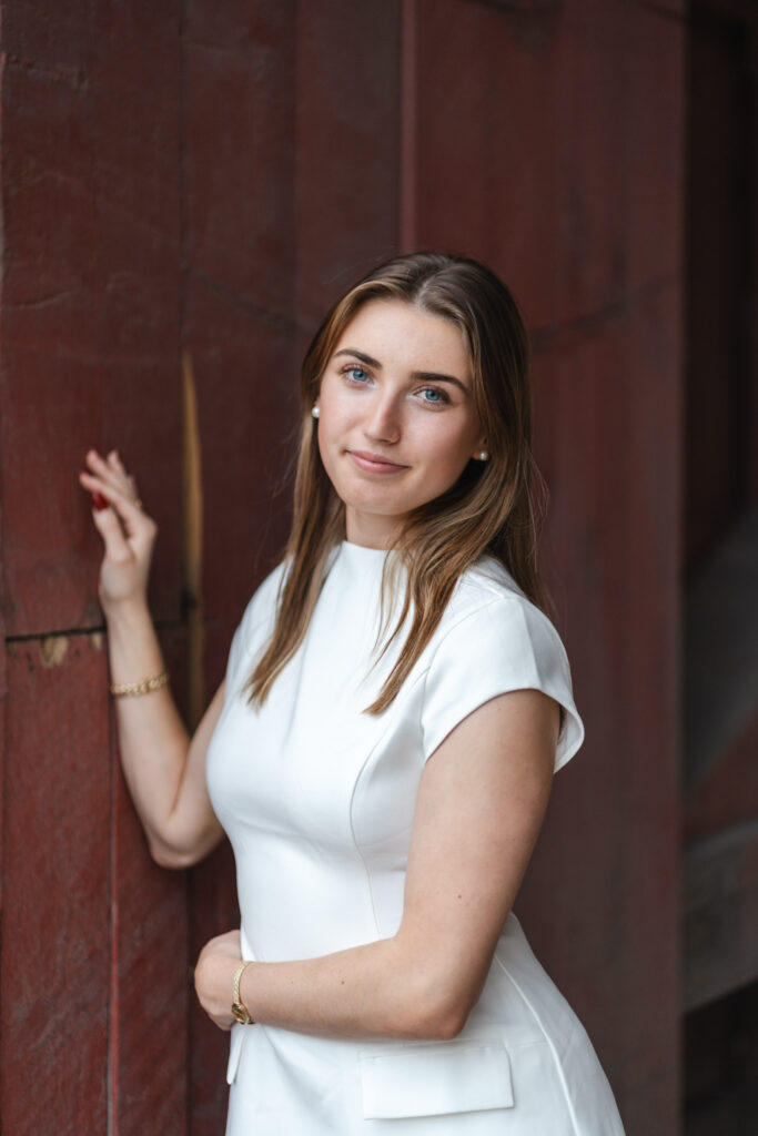 mid-length portrait of woman in white dress standing to the side of a covered bridge, leaning on the wall with arms wrapped around her