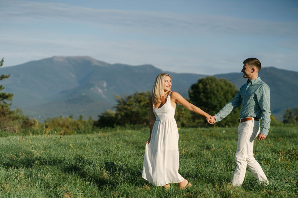 Couple walking across sunlit field with mountains in background, white mountains during engagement session