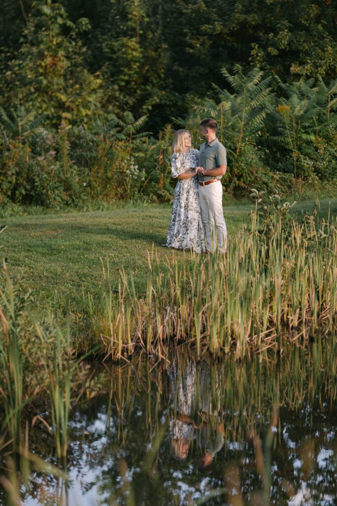 couple stand close and snuggle on the edge of sunlit pond